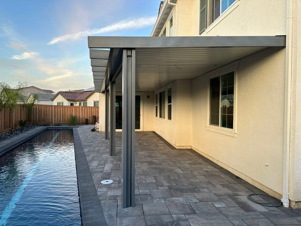 Modern backyard patio with a pool and shaded pergola, featuring stone flooring and scenic views.
