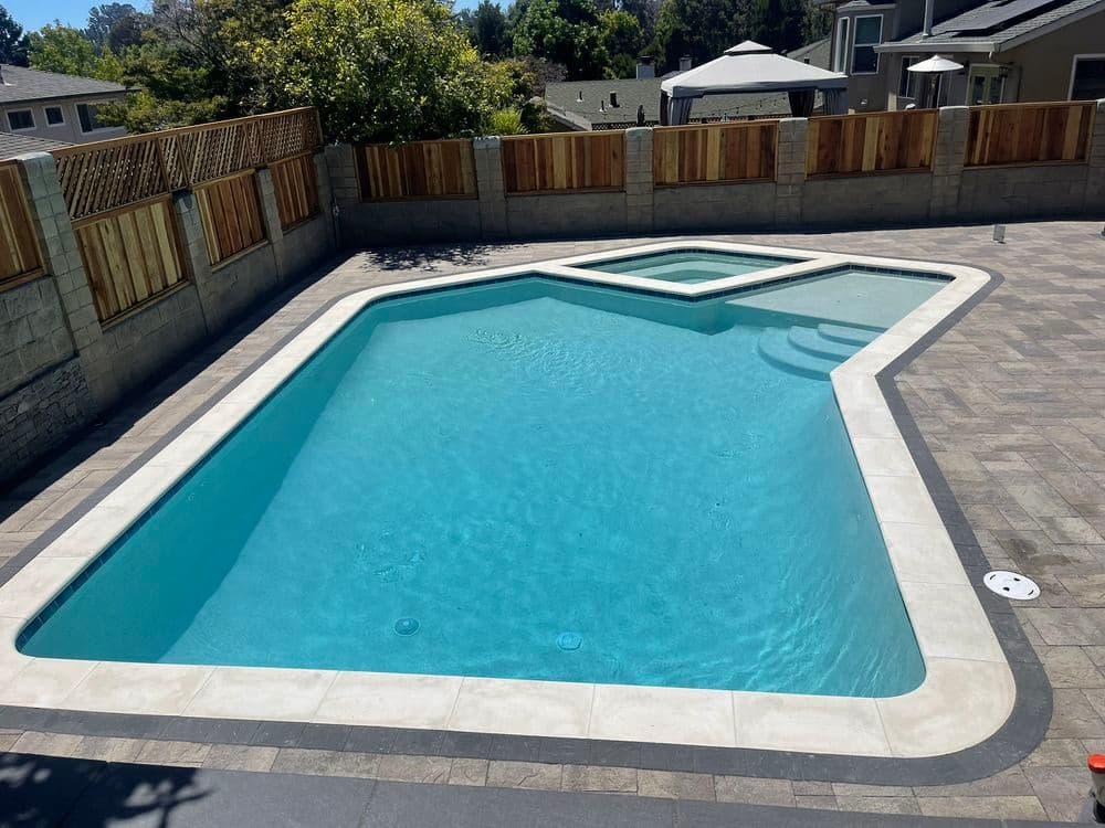 Modern outdoor swimming pool with a spa area, stone patio, and wooden fencing.