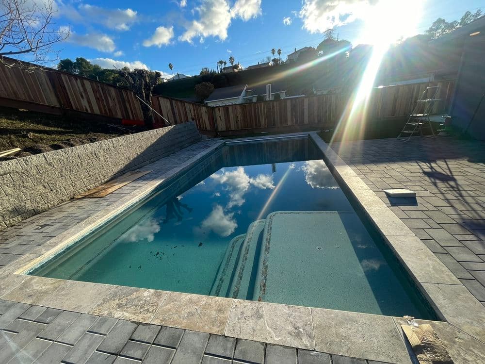 Modern residential swimming pool with clear water reflecting clouds, surrounded by stone patio.