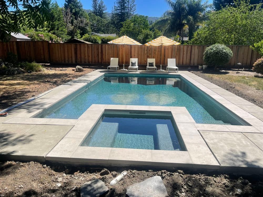 Modern backyard with a sparkling pool, spa, lounge chairs, and shaded areas under umbrellas.