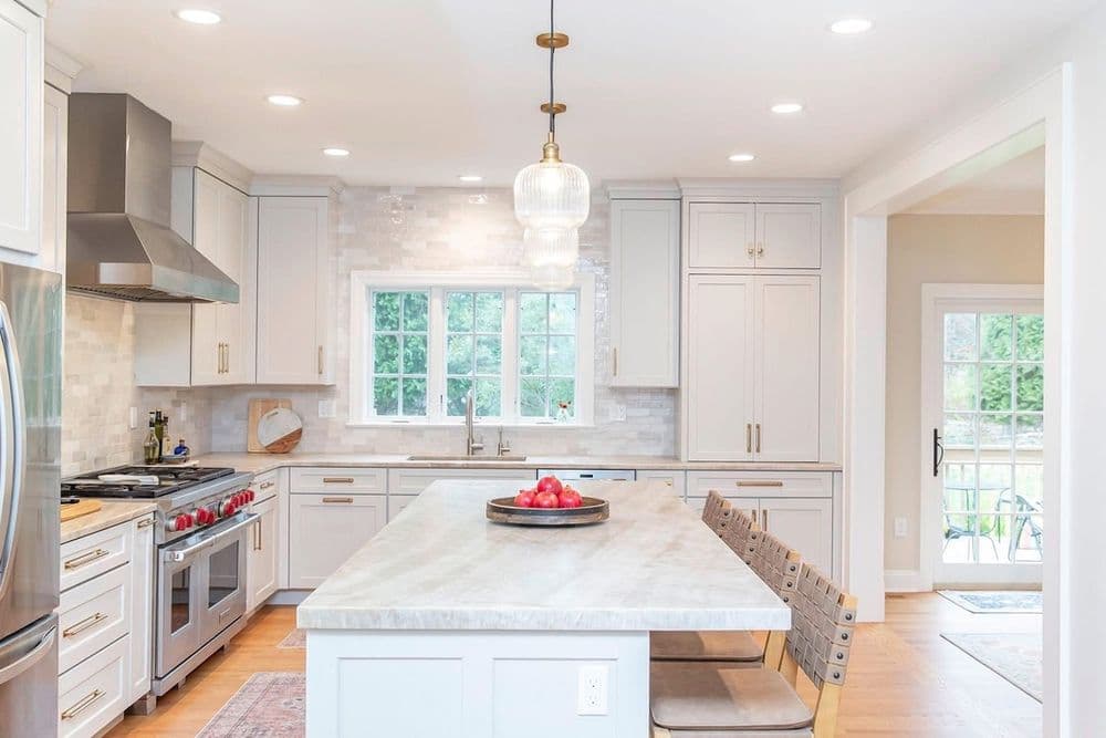 Modern kitchen with white cabinets, marble island, and stainless steel appliances, featuring natural light.
