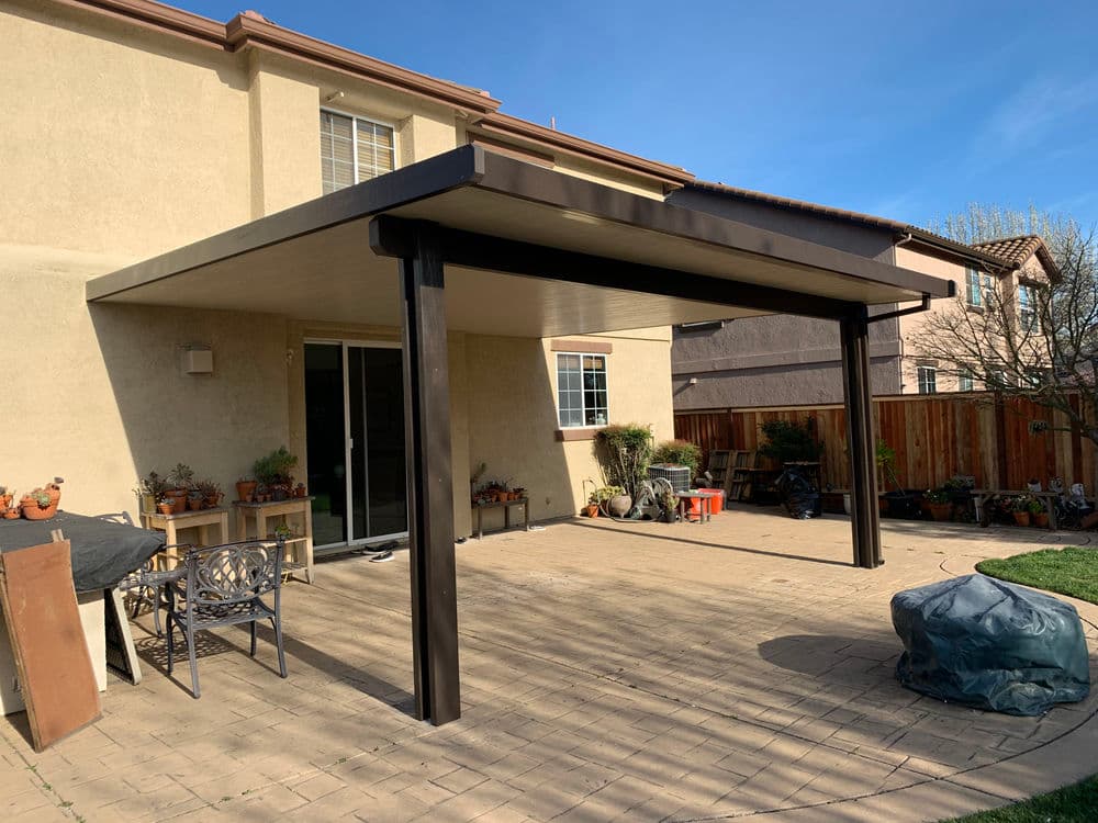 Backyard patio cover with dark metal frame and stone floor, surrounded by plants and furniture.