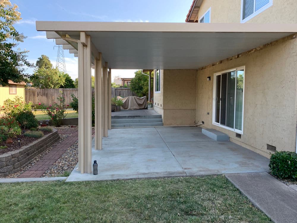 Patio cover over concrete slab in residential backyard with landscaping and window view.