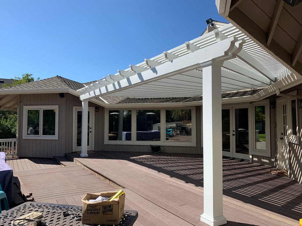 Patio with white pergola, surrounded by greenery and large glass windows. Sunny outdoor space.