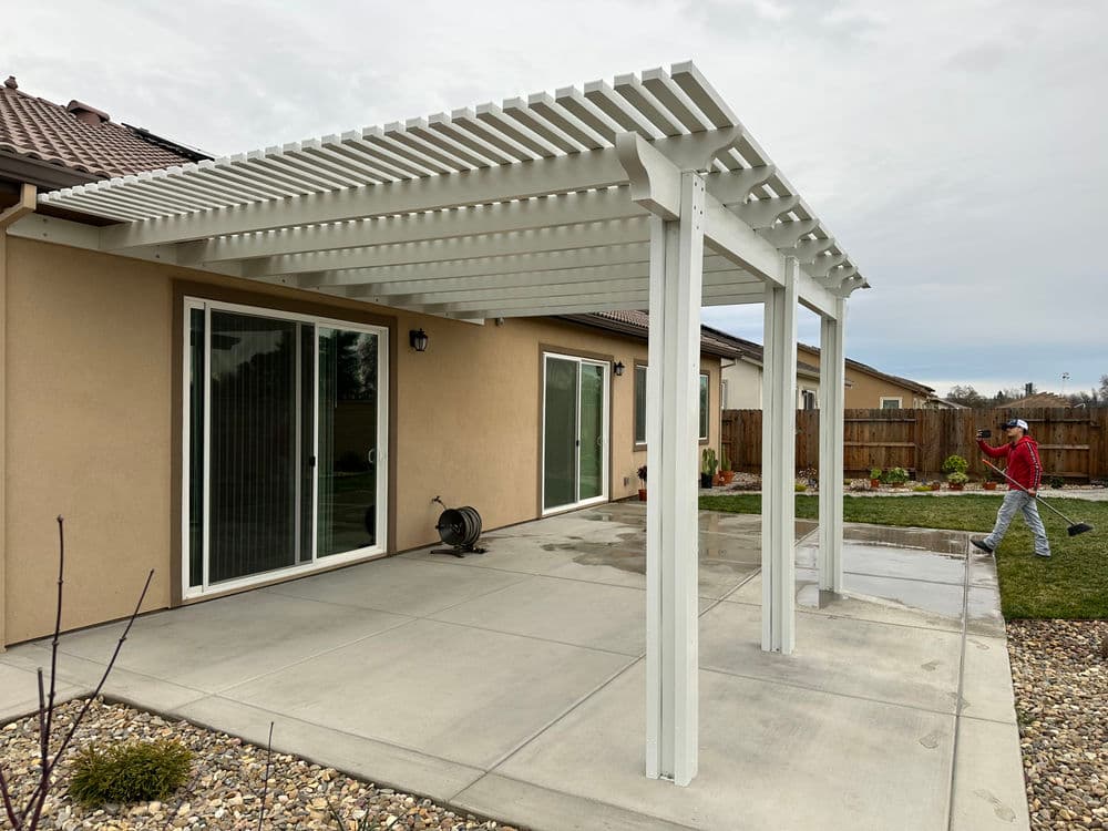 Modern white patio pergola beside a home with sliding glass doors and landscaped yard.