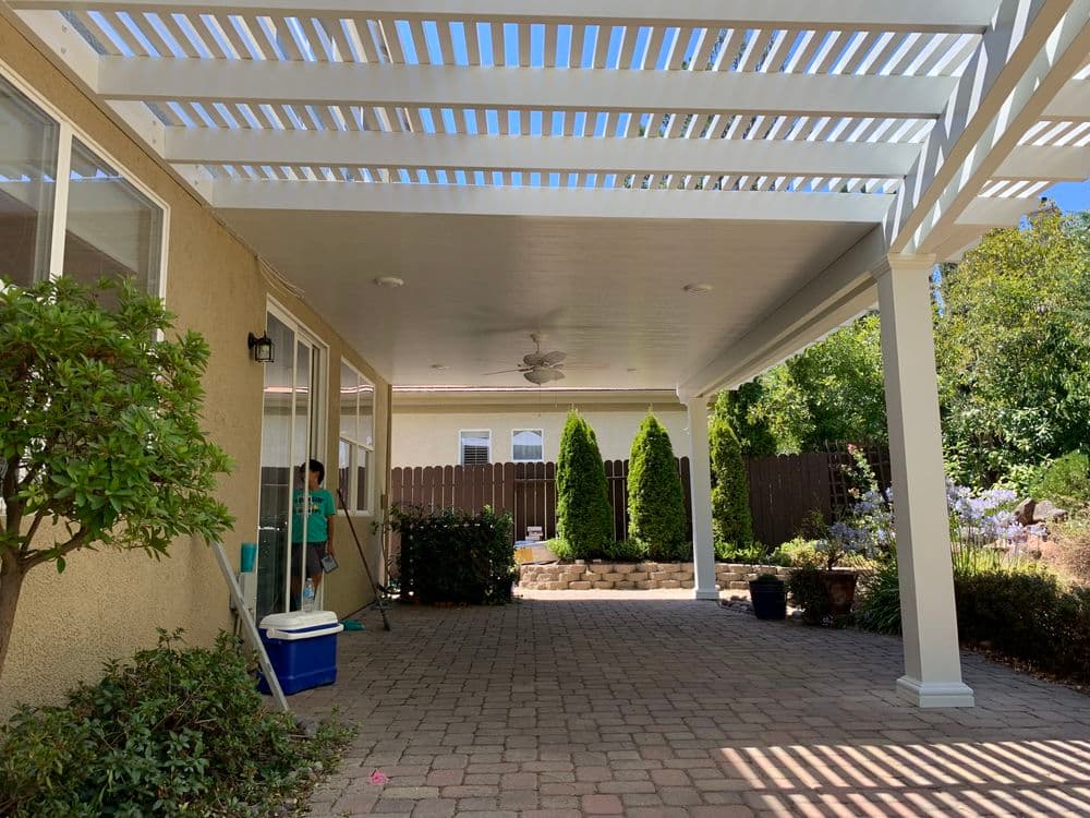 Outdoor patio with pergola, greenery, and a person cleaning near a cooler.