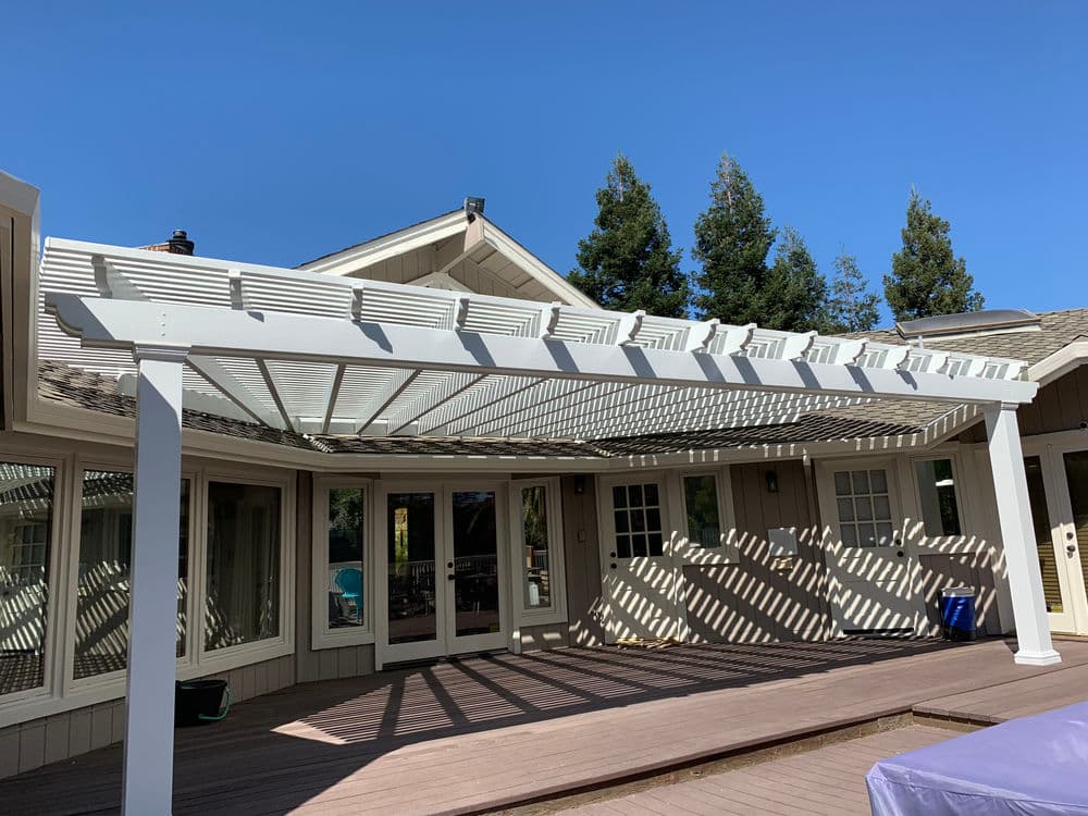 White pergola providing shade over a wooden deck in a sunny backyard with trees.