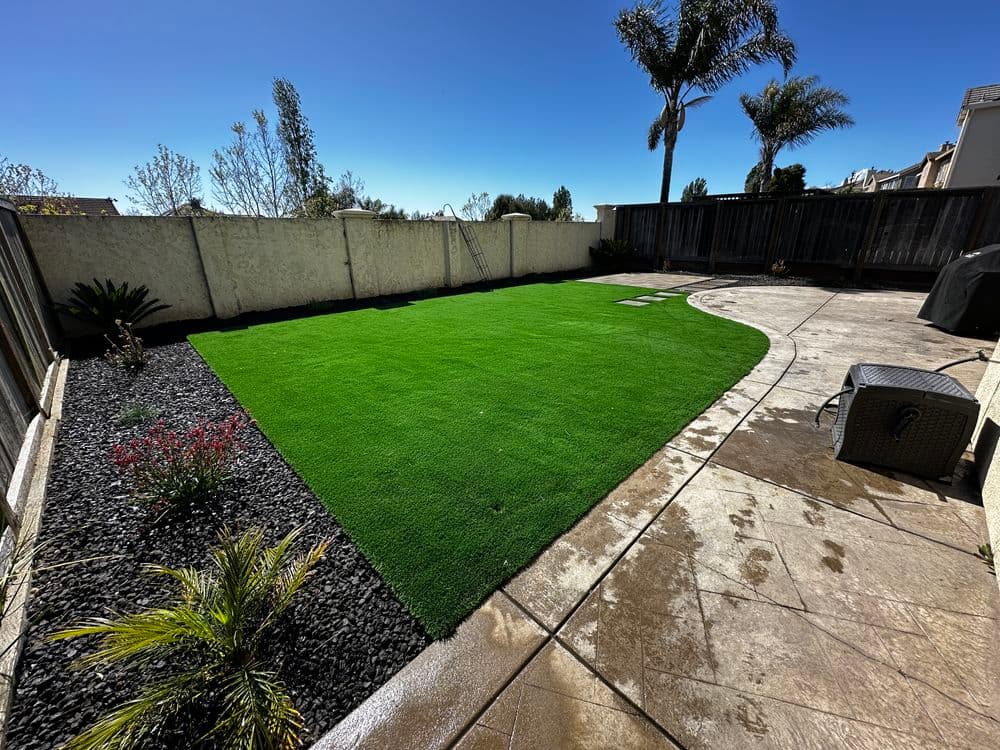 Lush green artificial grass yard with stone patio and landscaping features under clear blue sky.