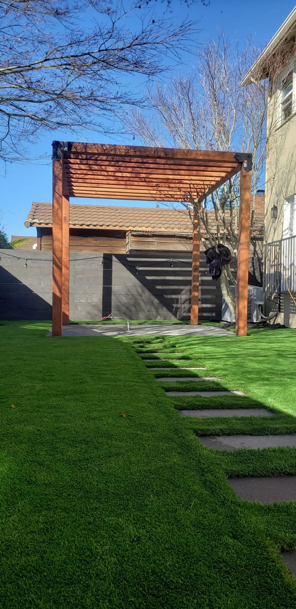 Wooden pergola in backyard with synthetic grass and stone pathway under clear blue sky.