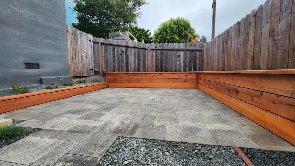 Modern outdoor patio with wooden seating, stone pavers, and fenced greenery area.