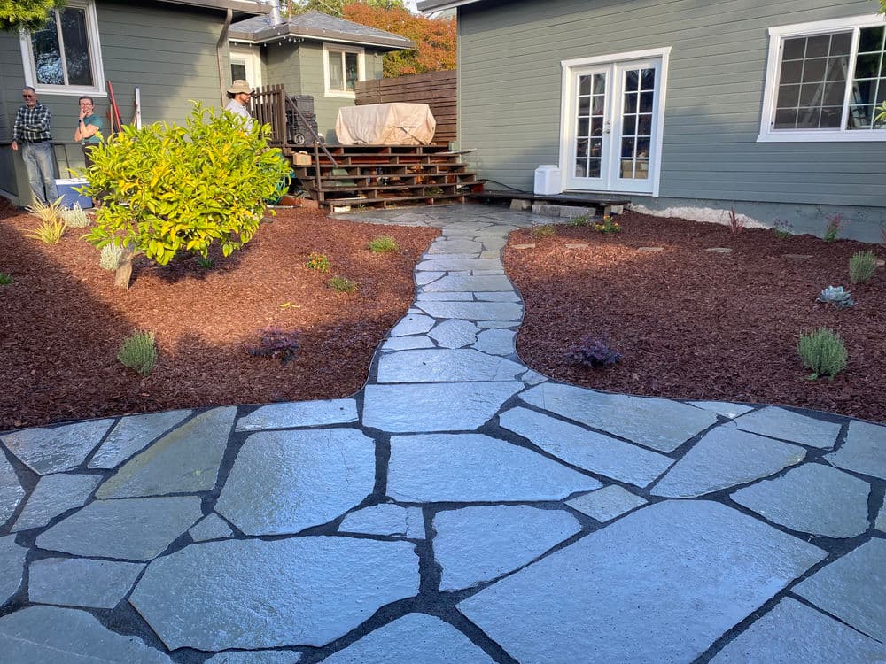 Flagstone pathway through landscaped yard with mulch, shrubs, and two houses in background.