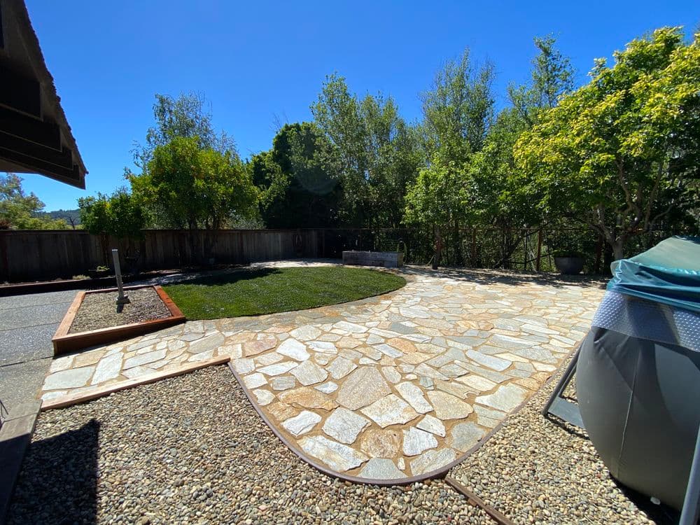Backyard landscape featuring stone patio, lush grass, trees, and hot tub under clear blue sky.