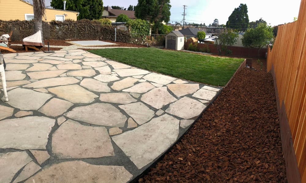Backyard landscape featuring stone patio, grassy area, and wood fence in residential setting.