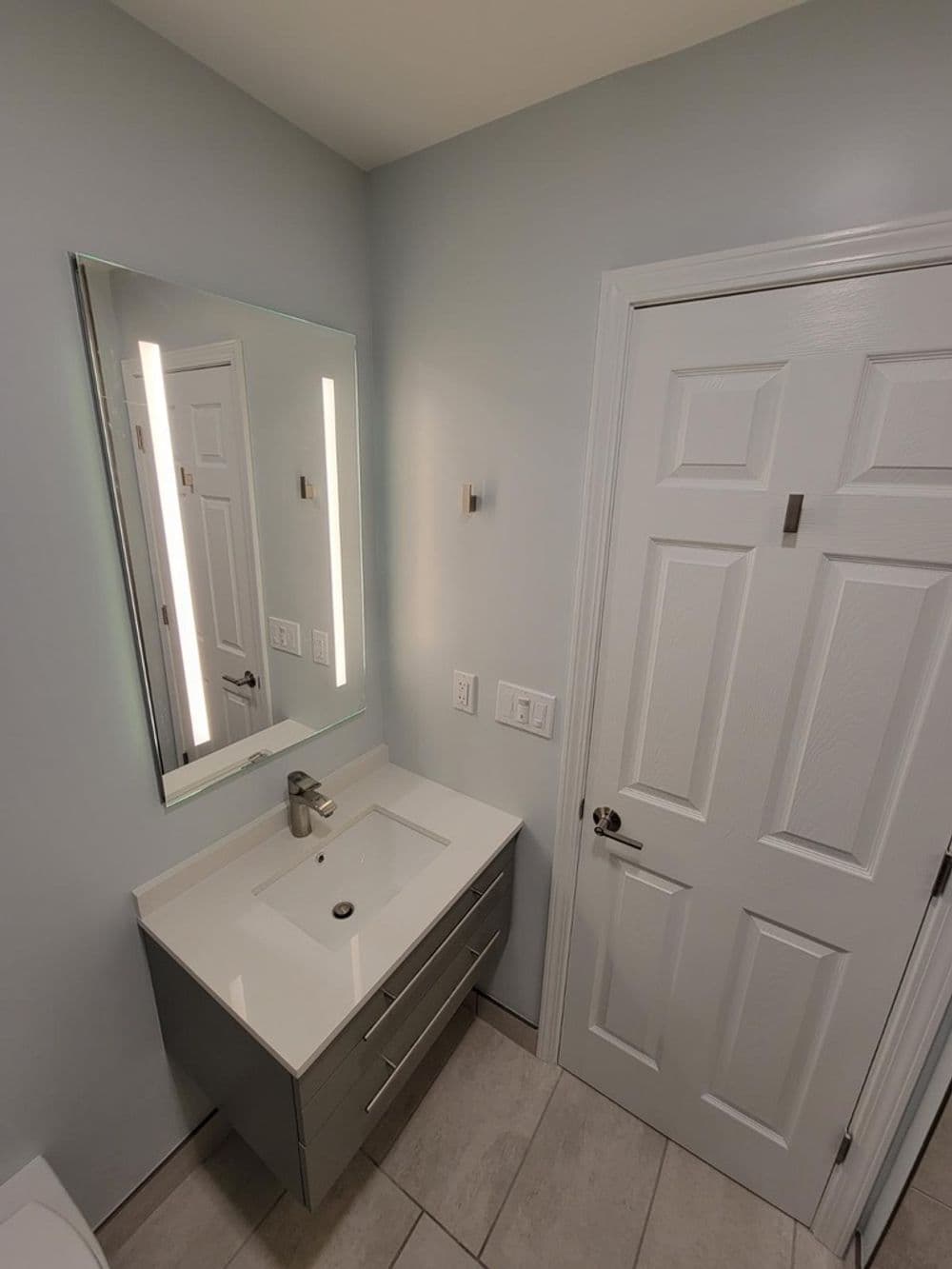 Modern bathroom with gray vanity, illuminated mirror, and white paneled door.