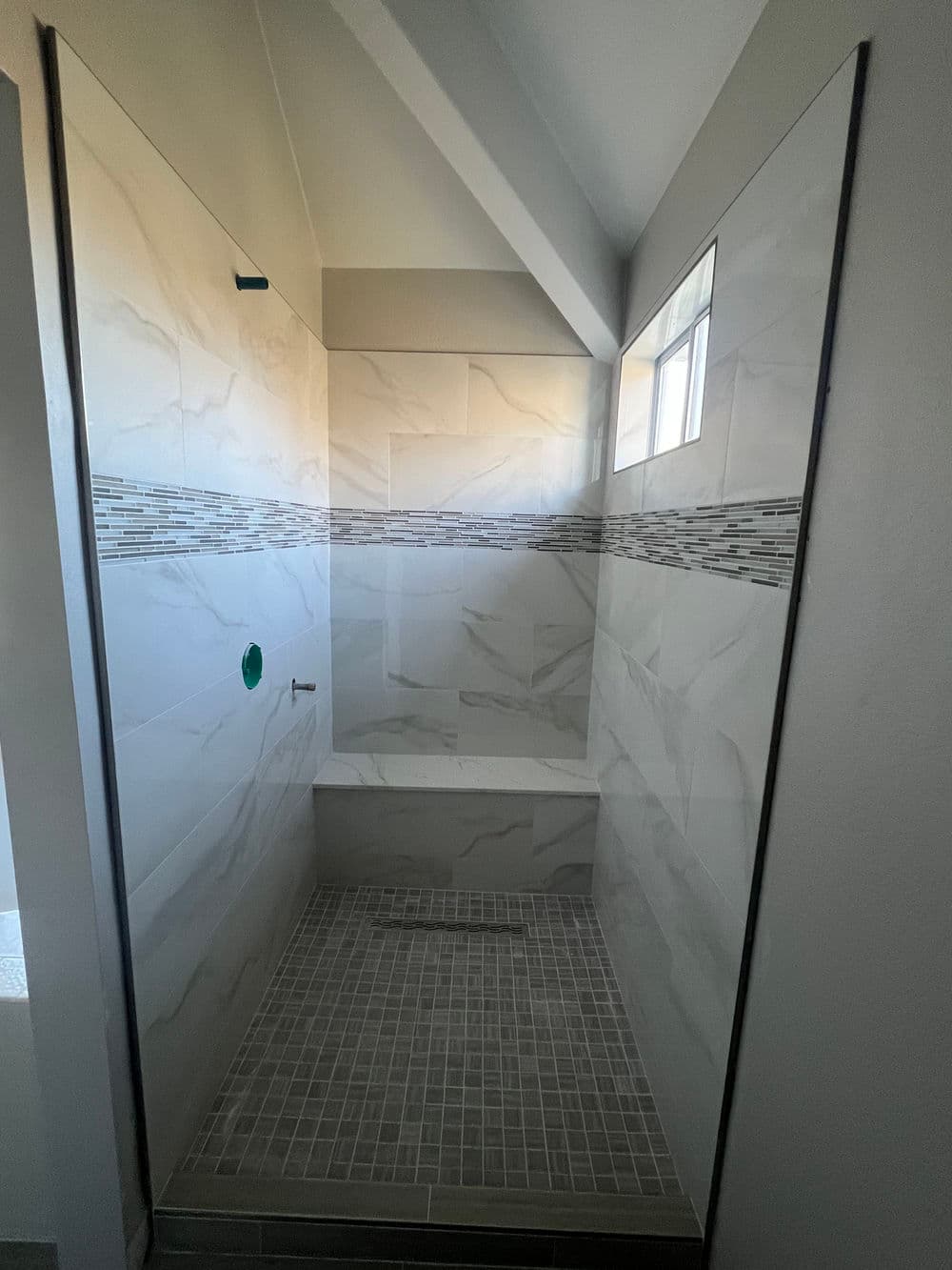 Modern shower with marble-like tiles, a built-in bench, and natural light from window.