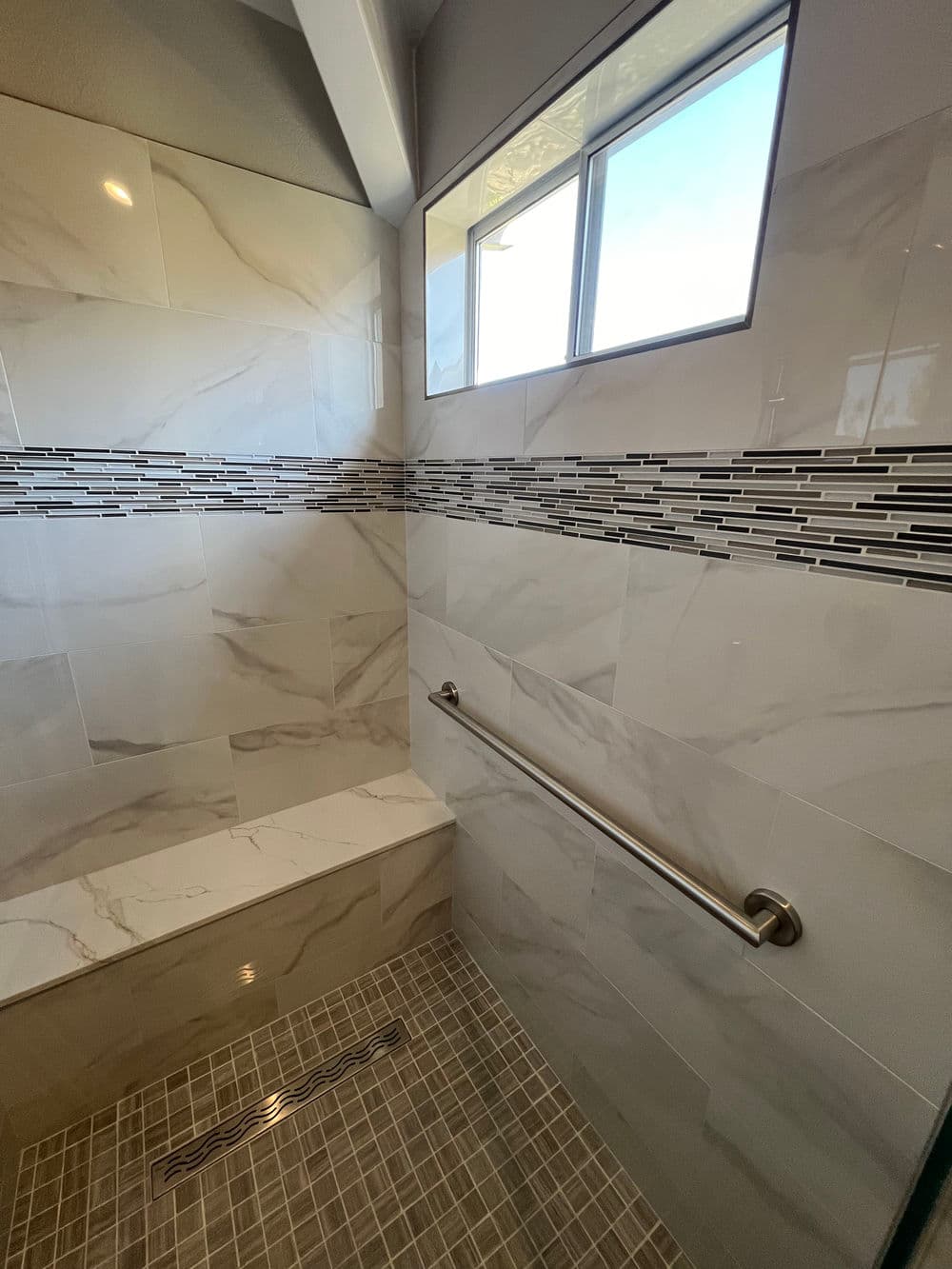 Modern shower featuring marble tiles, accent strip, grab bar, and natural light from window.